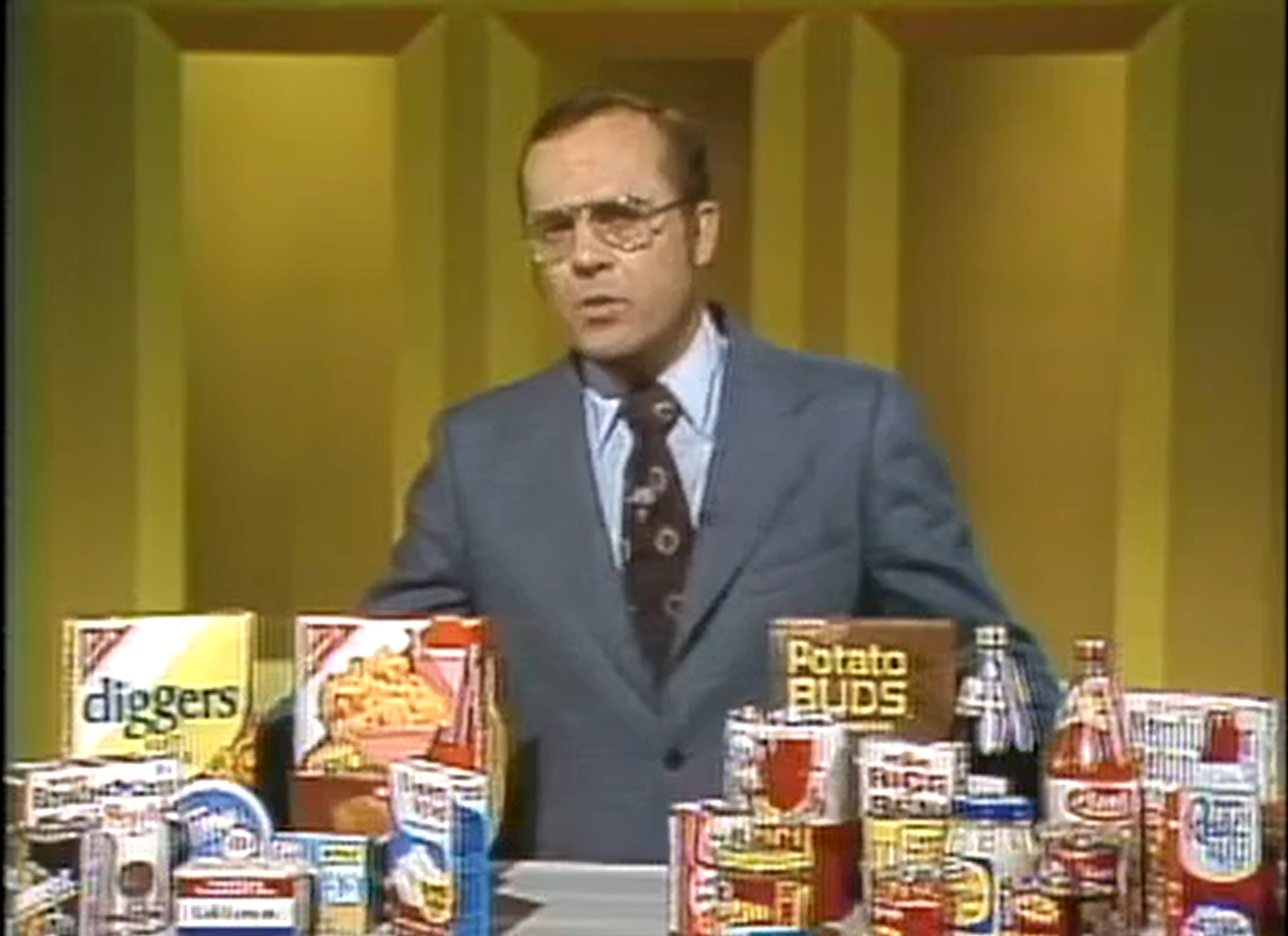 Male television presenter standing behind a table covered with consumer goods of varying types, including Reddi Whip, Potato Buds, and Diggers. 