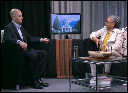 A white man seated across from a Black woman, both in black armchairs, with a television in the background that reads "Higher Education Today."