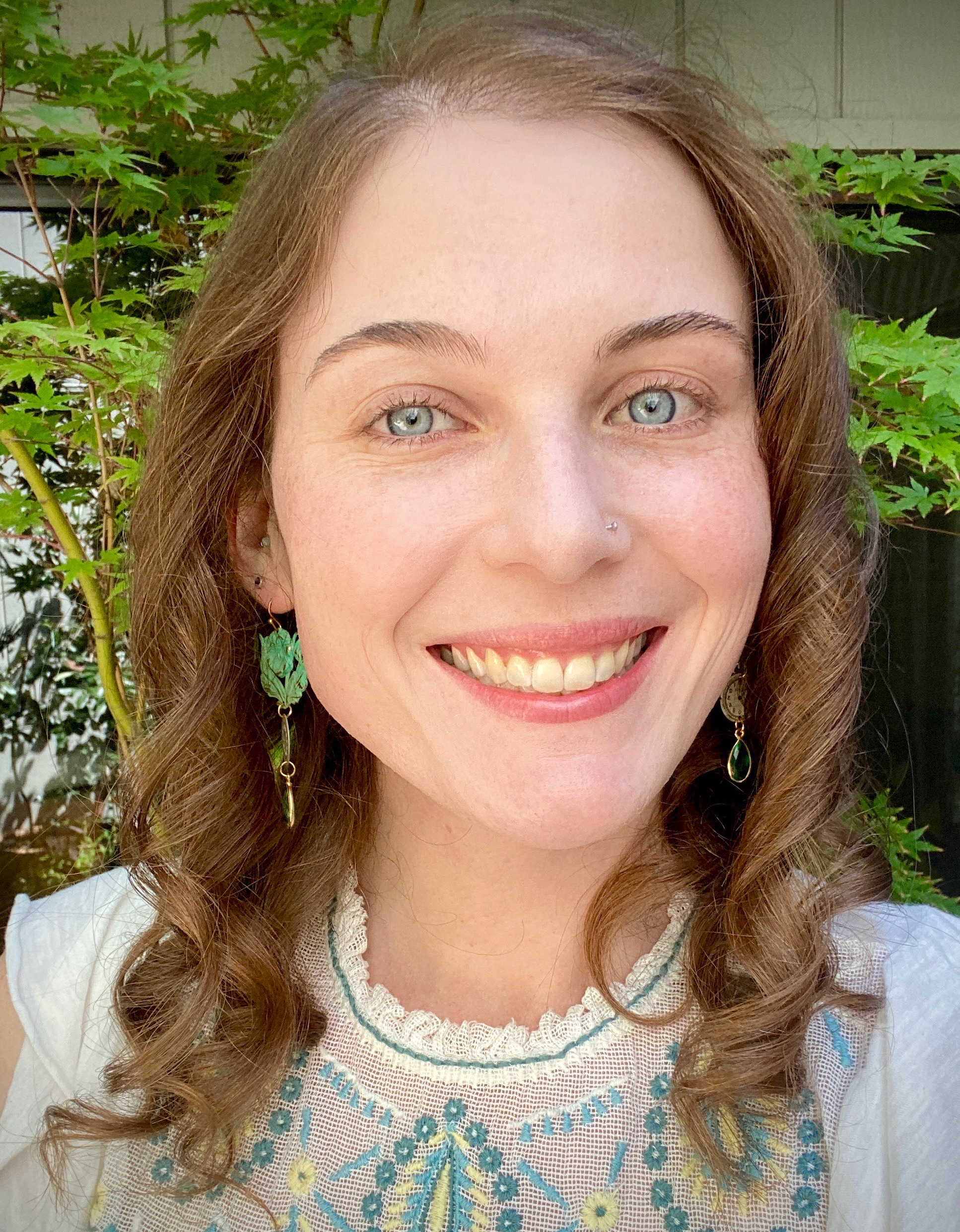 Headshot of Sabrina Gunn, a white woman with blue eyes and auburn hair. Sabrina is smiling and wearing a white blouse, green dangle earrings, and a nose piercing.